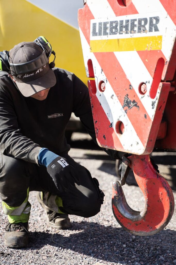 Mastering the First Impression: Your intriguing post title goes here A construction worker crouches to inspect a large machinery hook outdoors, wearing cap and headphones.